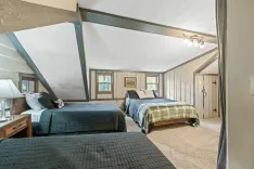 Bright and cozy attic bedroom interior with sloped ceilings, two beds with quilts, nightstands with lamps, framed artwork, and a carpeted floor.