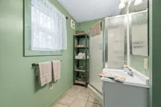 A small bathroom with green walls, white fixtures, a shower stall, and a window with a curtain.