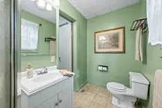Alt: A small bathroom with mint green walls, featuring a white vanity sink, a framed mirror with overhead lighting, a toilet, a tan tiled floor, and a framed picture on the wall.