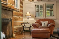 Cozy cabin interior with a plush leather armchair, wooden side table with a lamp, and part of a fireplace, against a rustic wooden wall with a window.