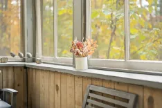 A cozy window nook with a wooden sill, a white vase with dried flowers, and a view of autumn foliage outside.