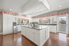 A spacious kitchen with white cabinetry, modern appliances, and an island, featuring a checkered and floral border wallpaper, hardwood floors, and natural light coming from large windows and glass doors leading to an outdoor deck.
