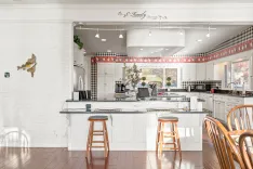 Bright and cozy kitchen interior with white cabinets, a central island with bar stools, decorative checkered wall trim, and sunlight streaming through the windows.