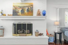 A cozy living room corner with a white-painted brick fireplace adorned with a decorative screen that says "Build a bridge and get over it", flanked by a wooden lantern on the left and a small lamp on a round table to the right. Above the mantel is a nautical painting of boats on water, beside decorative items including a string of wooden fish, a blue vase, and a pair of binoculars.