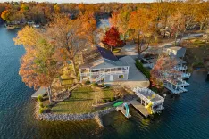 Aerial view of a lakeside house with autumn-colored trees, a dock, and boats during fall season.