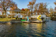 Lakeside house with a white porch, a yellow slide, and a green paddle boat on a sunny day with autumn trees.