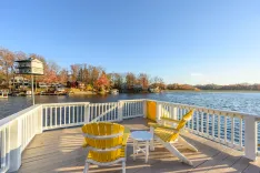 Lakeside wooden deck with two yellow Adirondack chairs and a white table, overlooking a calm lake with autumn-colored trees and houses in the background.