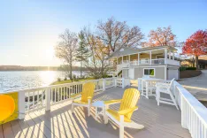 Lakeside house with a spacious deck, Adirondack chairs oriented towards the water, and autumn foliage in the background during sunset.