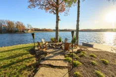 Stone pathway leading to a lakeside seating area with Adirondack chairs and a fire pit, with autumn trees and houses in the background, under a clear blue sky.