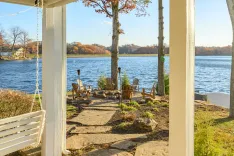 A serene lakeside setting with Adirondack chairs arranged around a fire pit, viewed from a porch with a swing, against a backdrop of autumn trees.