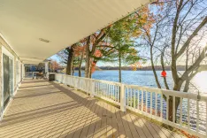 Spacious lakeside deck with outdoor furniture, surrounded by trees with autumn leaves, and a tranquil lake view under a clear blue sky.