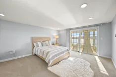 Bright and airy modern bedroom with a wooden bed frame, striped bedding, large shaggy area rug, and sliding glass doors leading to a balcony with a view of the water.