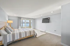 Spacious bedroom with a large bed, bedside table with a lamp, mounted TV, carpet flooring, and sliding glass doors leading to a balcony with a view of the water.