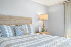 A cozy beach-themed bedroom with a distressed wooden headboard, blue and white striped bedding, nautical decorative pillows, and a bedside table with a lamp.