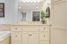 A well-lit bathroom interior with beige cabinets, a large mirror reflecting the closet in the bedroom, a potted plant on the countertop next to the sink, and a hanging towel.