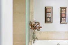 A serene bathroom corner with a cream-colored tile wall, a clear glass vase with eucalyptus branches on the edge of a bathtub, beside two framed pictures of leaves on the wall.