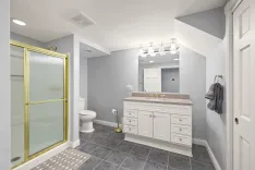 Modern bathroom interior with a glass-enclosed shower, vanity cabinet with sink, toilet, and towel on a hanger, featuring tiled floors and soft gray walls.