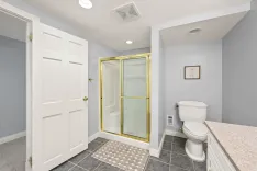 Modern bathroom interior with a closed white door, gray tiled floor, glass-enclosed shower with golden frame, white toilet, and a granite countertop against light blue walls with a decorative sign that reads "TAKE A SEAT".