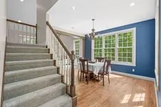 Elegant dining room with a blue accent wall, large windows, and a wooden table set adjacent to a staircase with white spindles and wooden banister.