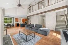 Bright and airy open-concept living space with a sectional sofa, hardwood floors, high ceiling with a fan, and an adjoining kitchen with wooden cabinets.