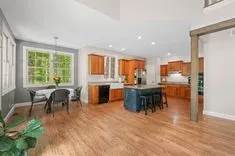 Spacious kitchen with wooden floor and cabinets, black appliances, and a small dining area with a view of greenery outside the windows.