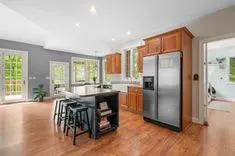 Spacious modern kitchen with wooden cabinets, stainless steel appliances, central island with stools, and hardwood flooring, leading to a deck through French doors.