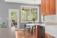 Bright kitchen with gray walls showcasing wooden cabinets and a dining area with a white tablecloth and a bouquet of flowers, leading to a sunlit porch through French doors.