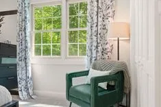Brightly lit room with a green armchair, throw blanket, floor lamp, leaf-patterned curtains, and a view of greenery outside the window.