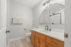 Modern bathroom interior with wooden vanity cabinet, white countertop with sink, large round mirror, and lighting fixtures above.