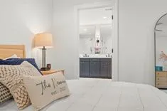Cozy bedroom interior with a view into a modern bathroom, featuring a bed with decorative pillows, lamp on a nightstand, and a framed wall art.