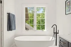 Bright and modern bathroom with a freestanding white bathtub, black faucet, towel hanging on the wall, and a window overlooking green trees.