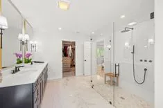 Modern bathroom interior with dual vanity, marble floor, glass shower, and walk-in closet in the background.