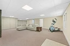 Spacious basement room with a sectional sofa, exercise bike, tiled entryway, and fluorescent lighting.