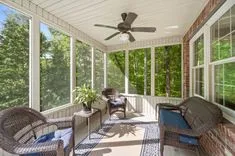 Bright sunroom with wicker furniture, ceiling fan, and large windows overlooking a lush forest.