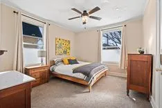 Cozy bedroom with a queen-sized bed, two windows with blinds, a ceiling fan, and a nightstand.