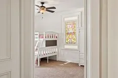 View from a doorway of a cozy bedroom with a white toddler bed, a ceiling fan, and a stained glass window.