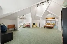Interior of a spacious attic room with slanted ceilings, equipped with various furniture including a bed, chairs, and a bench, and lit by track lighting.