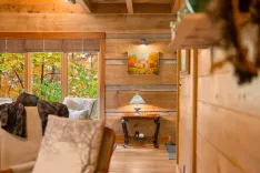 Cozy wooden cabin interior with large windows showing autumn foliage outside, decorative table with lamp, and comfortable seating area.
