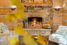 Cozy rustic living room with a stone fireplace lit with a crackling fire, decorated mantle with antlers and pumpkins, and comfortable furnishings with warm accents.