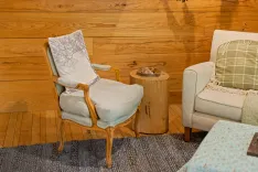 A cozy sitting area with two upholstered chairs, one with a tree-print pillow, a wooden stump side table with a tray and mugs, all on a textured rug against a wooden plank wall.