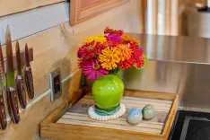 A vibrant bouquet of red, orange, yellow, and purple flowers in a green vase on a kitchen counter, next to a knife block and decorative salt and pepper shakers.