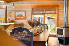 Cozy wooden cabin interior featuring a dining table decorated with mini pumpkins and dried plants, elegant blue chairs, a fireplace, and a fruit painting on the wall.