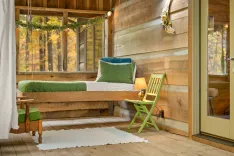 Cozy cabin interior with a wooden daybed, green chair, table lamp, and large windows overlooking autumn trees.