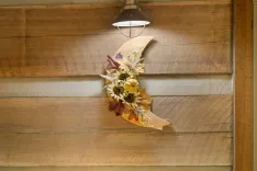 Alt: A pendant light illuminates a decorative arrangement of dried flowers and leaves on a wooden wall.