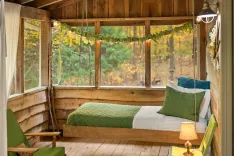 Cozy wooden cabin bedroom with a green and white bed, hanging greenery, a lamp, and a view of autumn trees through the window.