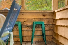 Two teal-colored metal bar stools against a wooden counter in a rustic porch setting with forest views visible through a window.