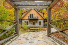 Rustic wooden cabin with a balcony framed by an open gate and surrounded by autumn-colored trees with a stone pathway leading to the house.