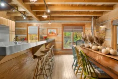 Rustic-style kitchen interior with wooden beams, a long dining table with green chairs, and a kitchen counter with bar stools. Decor includes pumpkins and dried plants, with natural light coming from the windows.