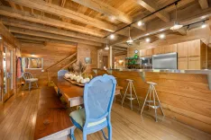Rustic wooden interior of a kitchen and dining area with stainless steel appliances, a long dining table with chairs, and a staircase in the background.