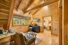 Cozy wooden cabin interior with warm lighting, exposed beams, a comfortable lounge area, bookshelf, and a glimpse into the bathroom.